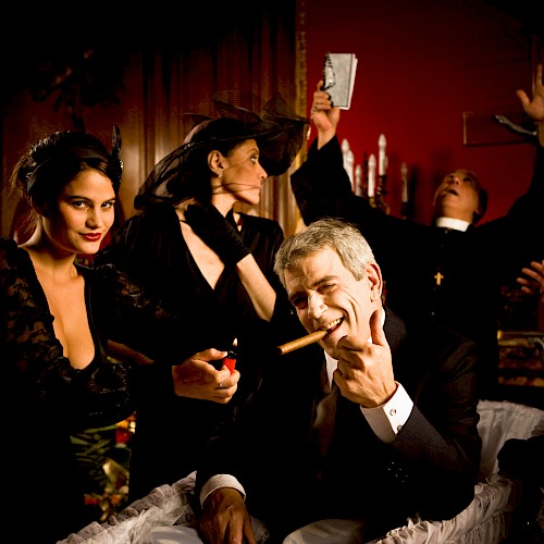 A man in a coffin with a cigar, surrounded by people, some in dramatic poses, in a dimly lit room.