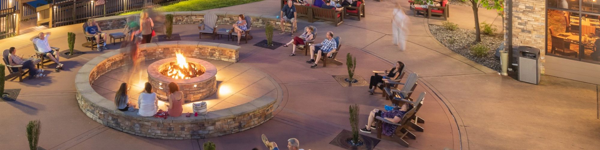 A scenic outdoor area with people sitting around a fire pit near a lake, surrounded by trees and string lights, next to a building with lights.