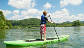 A person on a paddleboard in a lake, wearing a blue life vest. The background shows a forested hillside and blue sky.